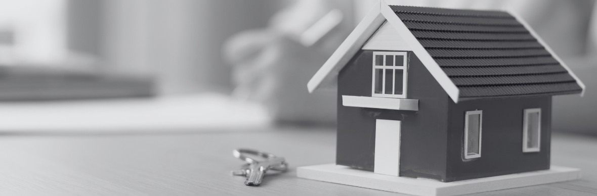 Close up of miniature model of a house with a key beside it. Out of focus background of person writing in the background.